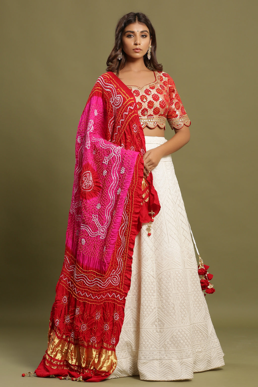 Shop White self-embroidered Zari lehenga set featuring bandhej dual print dupatta and zari & gota patti embroidered red blouse. The lehenga also has a long tassel attached to tie the knot. Pair it with beautiful jewelry to enhance your look. Shop online from Pure Elegance.