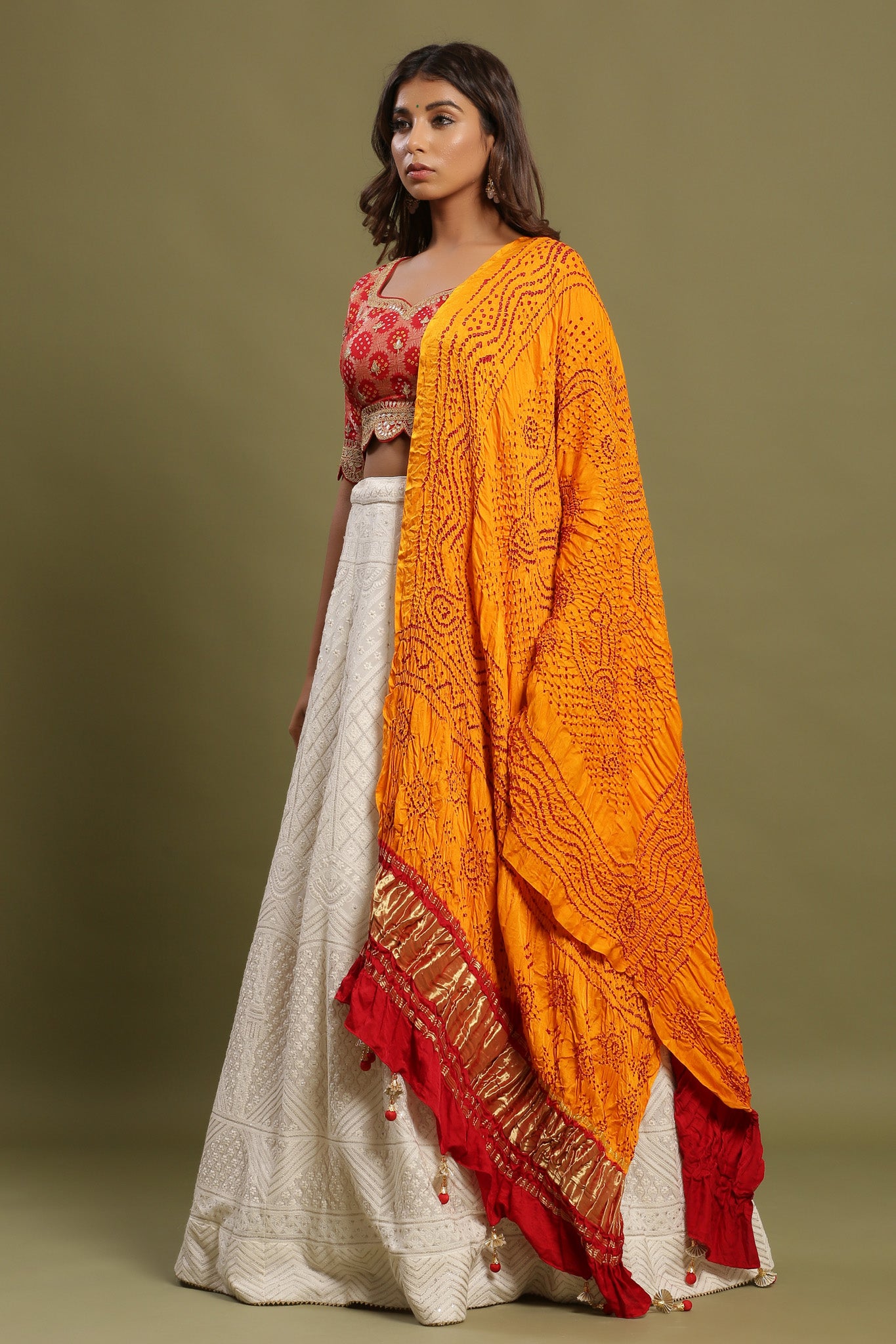 Shop White self-embroidered Zari lehenga set featuring bandhej orange print dupatta and zari & gota patti embroidered red balouse. The lehenga also has a long tassel attached to tie the knot. Pair it with beautiful jewelry to enhance your look. Shop online from Pure Elegance.